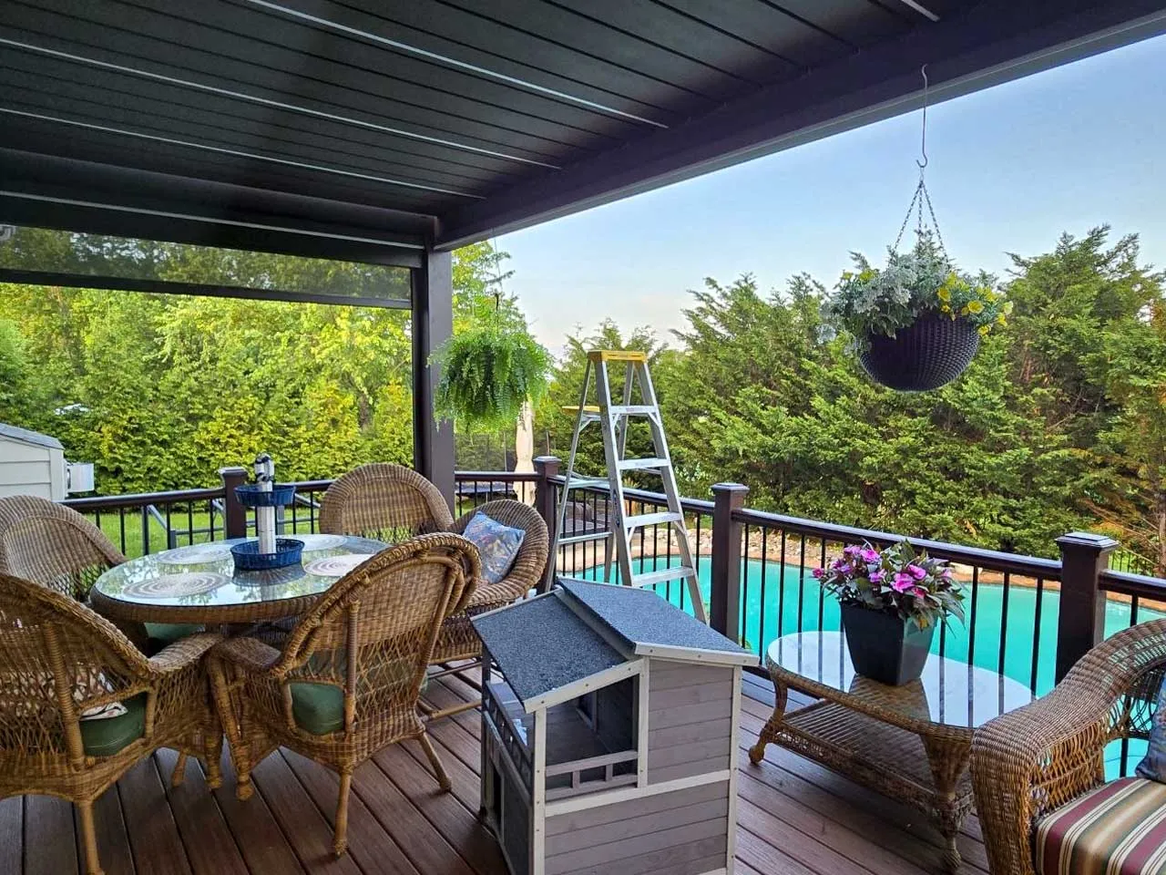 Dining set on a deck under a dark louvered pergola with a side screen, pool and trees beyond